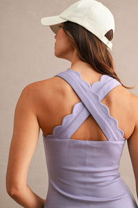 Woman wearing a lavender athelisure mini dress that has a scalloped, square neckline with a mini-length hem that also has a scalloped design indoors against a neutral backdrop. The photo shows the back of the dress and how that straps criss cross in the back. 