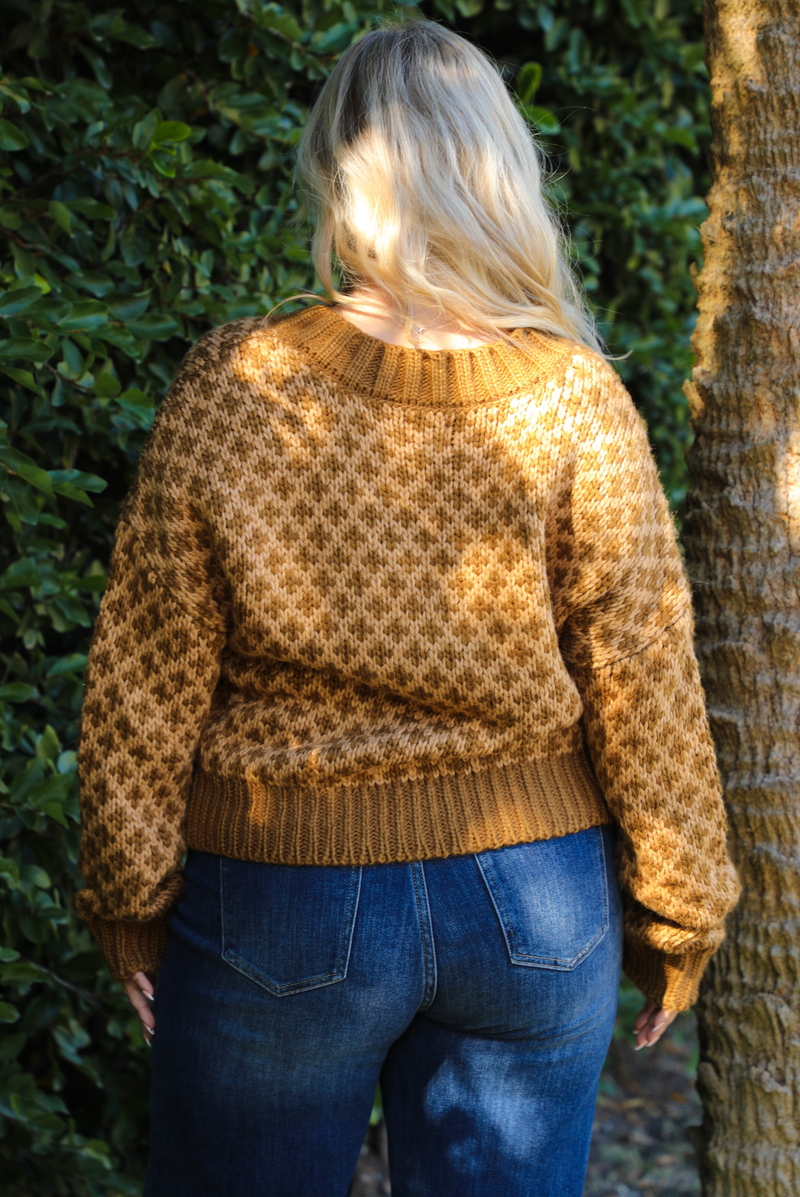 The back of a woman in a brown and tan patterned sweater and blue jeans leaning against a tree with greenery in the background.