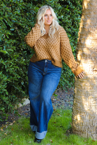 Woman in a brown and tan patterned sweater and blue jeans leaning against a tree with greenery in the background.
