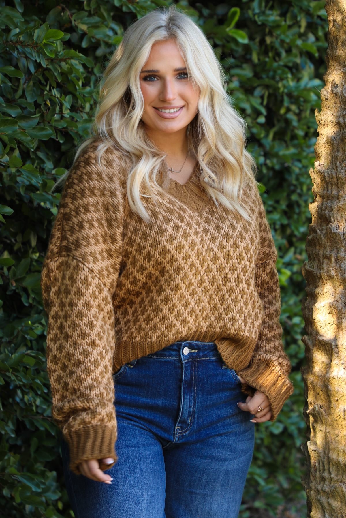 Woman in a brown and tan patterned sweater and blue jeans leaning against a tree with greenery in the background.