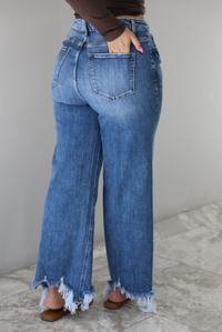 Woman wearing a medium wash denim that are high waisted, have functional front and back pockets, functional button and zipper closures, and have a raw, distressed hem. The photo shows the back of jeans, and the models hand is in the back right pocket. The photo is taken indoors against a white wall with white marble flooring.