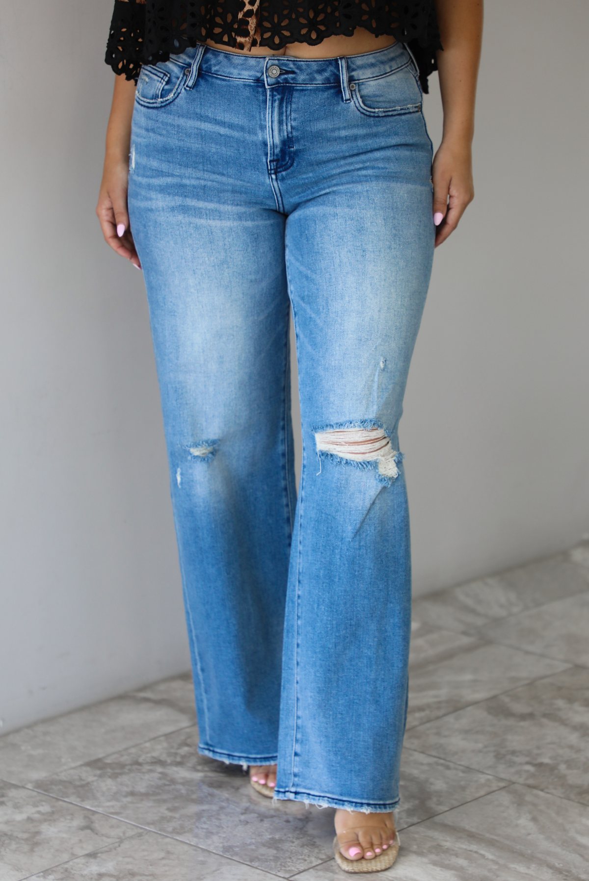 Woman wearing a pair of medium-wash, mid-rise denim jeans that have distressed details in the front and a flared, boot-cut-like hem. The photo is taken indoors against a white wall with white marble flooring. 