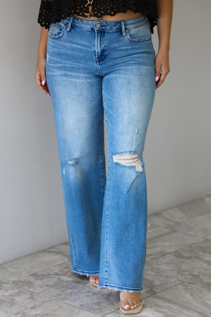 Woman wearing a pair of medium-wash, mid-rise denim jeans that have distressed details in the front and a flared, boot-cut-like hem. The photo is taken indoors against a white wall with white marble flooring. 