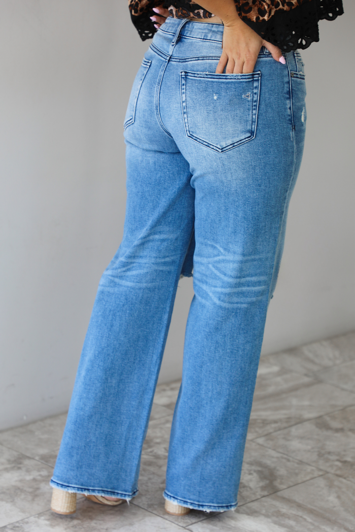 Woman wearing a pair of medium-wash, mid-rise denim jeans that have distressed details in the front and a flared, boot-cut-like hem. The photo is taken indoors against a white wall with white marble flooring and shows the back of the jeans. 