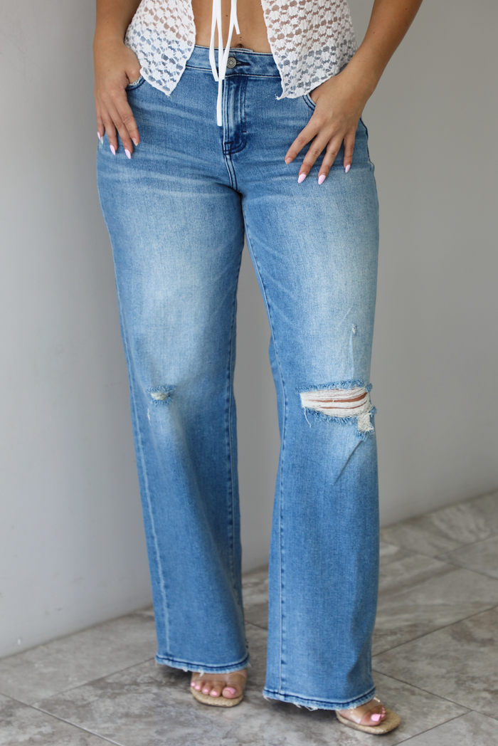 Woman wearing a pair of medium-wash, mid-rise denim jeans that have distressed details in the front and a flared, boot-cut-like hem. The photo is taken indoors against a white wall with white marble flooring. 