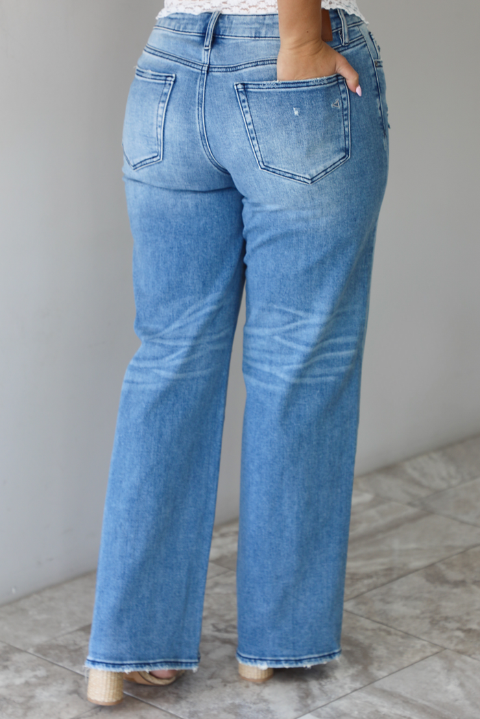 Woman wearing a pair of medium-wash, mid-rise denim jeans that have distressed details in the front and a flared, boot-cut-like hem. The photo is taken indoors against a white wall with white marble flooring and shows the back of the jeans. 