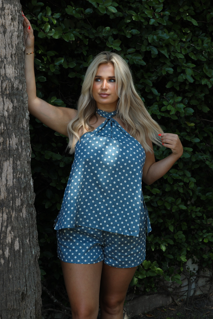 Woman wearing denim blue with white polka dot halter tie top and high waisted shorts outdoors with greenery in the background.