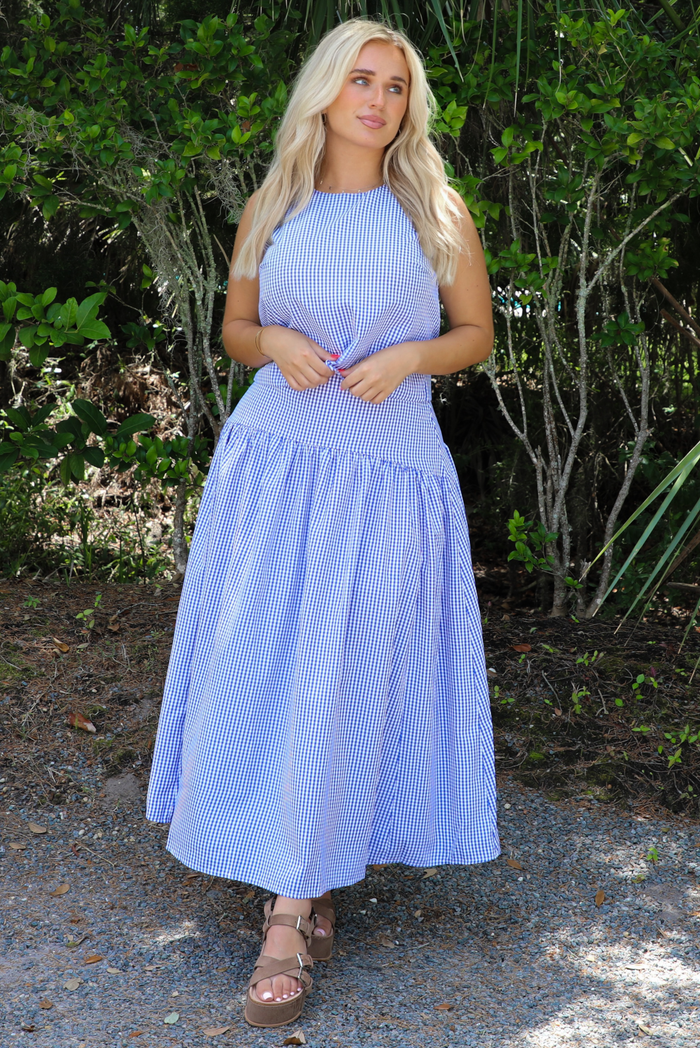 Woman wearing a blue and white gingham two piece set featuring a sleeveless top and high waisted, maxi length skirt outdoors with greenery in the background.