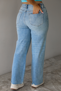 Woman wearing a pair of light-wash mid-rise denim jeans that has slight distressing with a raw, unfinished hem indoors against  a white wall with white marble flooring. The model is turned away with a hand in her back pockets showing the back of the denim. 