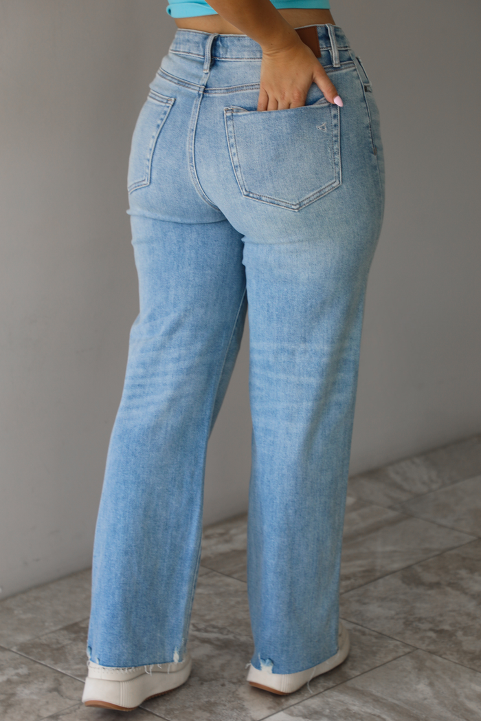 Woman wearing a pair of light-wash mid-rise denim jeans that has slight distressing with a raw, unfinished hem indoors against  a white wall with white marble flooring. The model is turned away with a hand in her back pockets showing the back of the denim. 