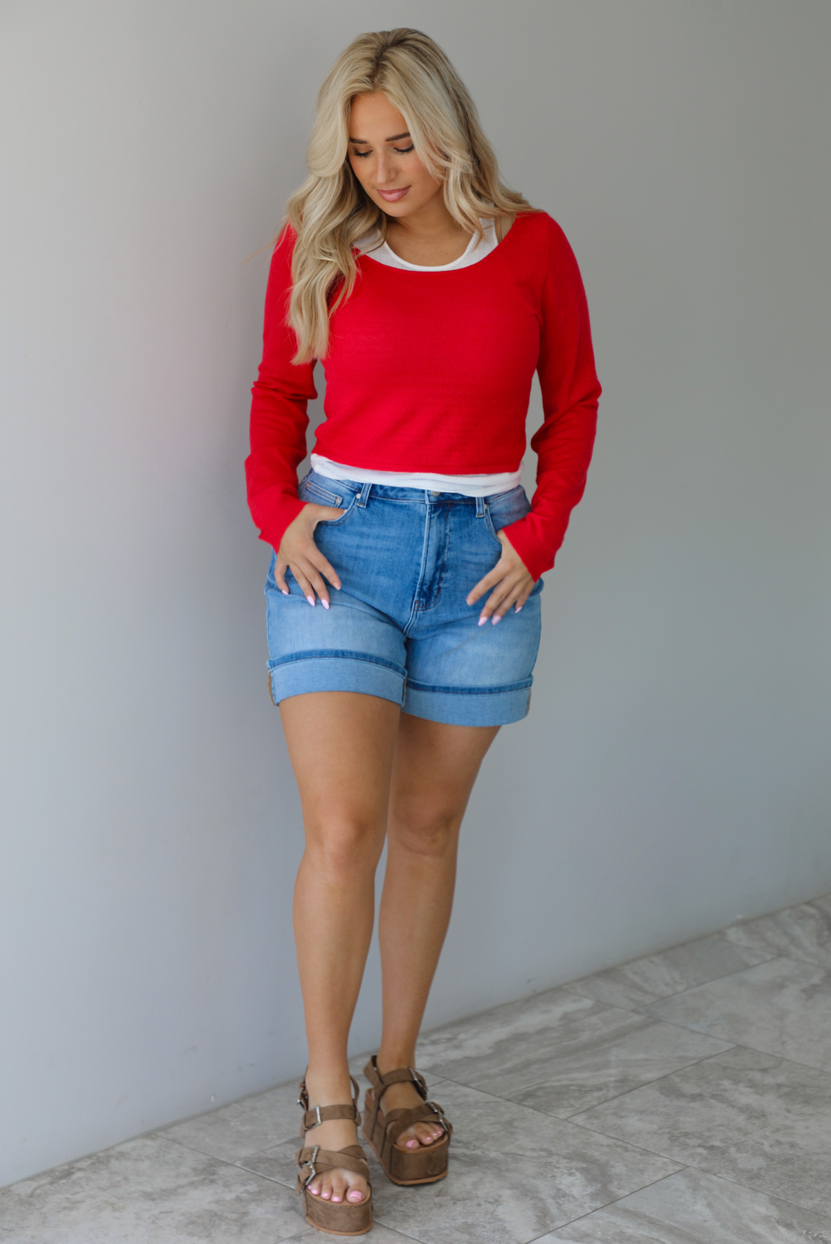 Woman wearing a red, long sleeved knit top with a white tank underneath with medium wash denim shorts indoors against a white wall with white marble flooring. 