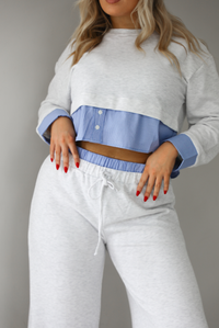 Woman wearing a heather grey two piece set with blue and white pin striped detailing at the hem of the top, at the wrists on the top, and at the waistline on the pants. The photo shows a closeup of the top and upper portion of the pants. The photo is taken indoors against a white wall with white marble flooring. 