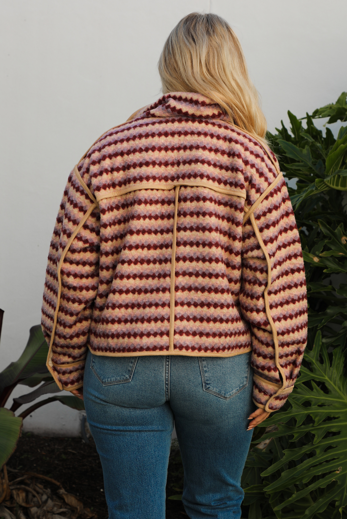 Jacket with a multi-colored sideways checkered pattern throughout, collared neckline, functional pockets in the front, and a functional zipper closure in the font. The jacket is paired with medium wash denim jeans and tan colored uggs. The photo is taken outdoors against a white wall with greenery in the back. The photo showcases the back of the jacket close up.
