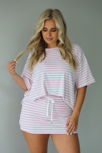 Woman wearing a pink and white striped two piece set featuring an oversized two and fitted mini skirt indoors against a white wall. 