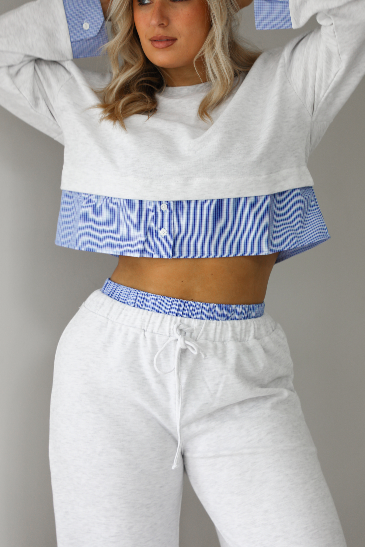 Woman wearing a heather grey two piece set with blue and white pin striped detailing at the hem of the top, at the wrists on the top, and at the waistline on the pants. The photo shows a closeup of the top and upper portion of the pants. The photo is taken indoors against a white wall with white marble flooring. 