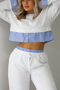 Woman wearing a heather grey two piece set with blue and white pin striped detailing at the hem of the top, at the wrists on the top, and at the waistline on the pants. The photo shows a closeup of the top and upper portion of the pants. The photo is taken indoors against a white wall with white marble flooring. 