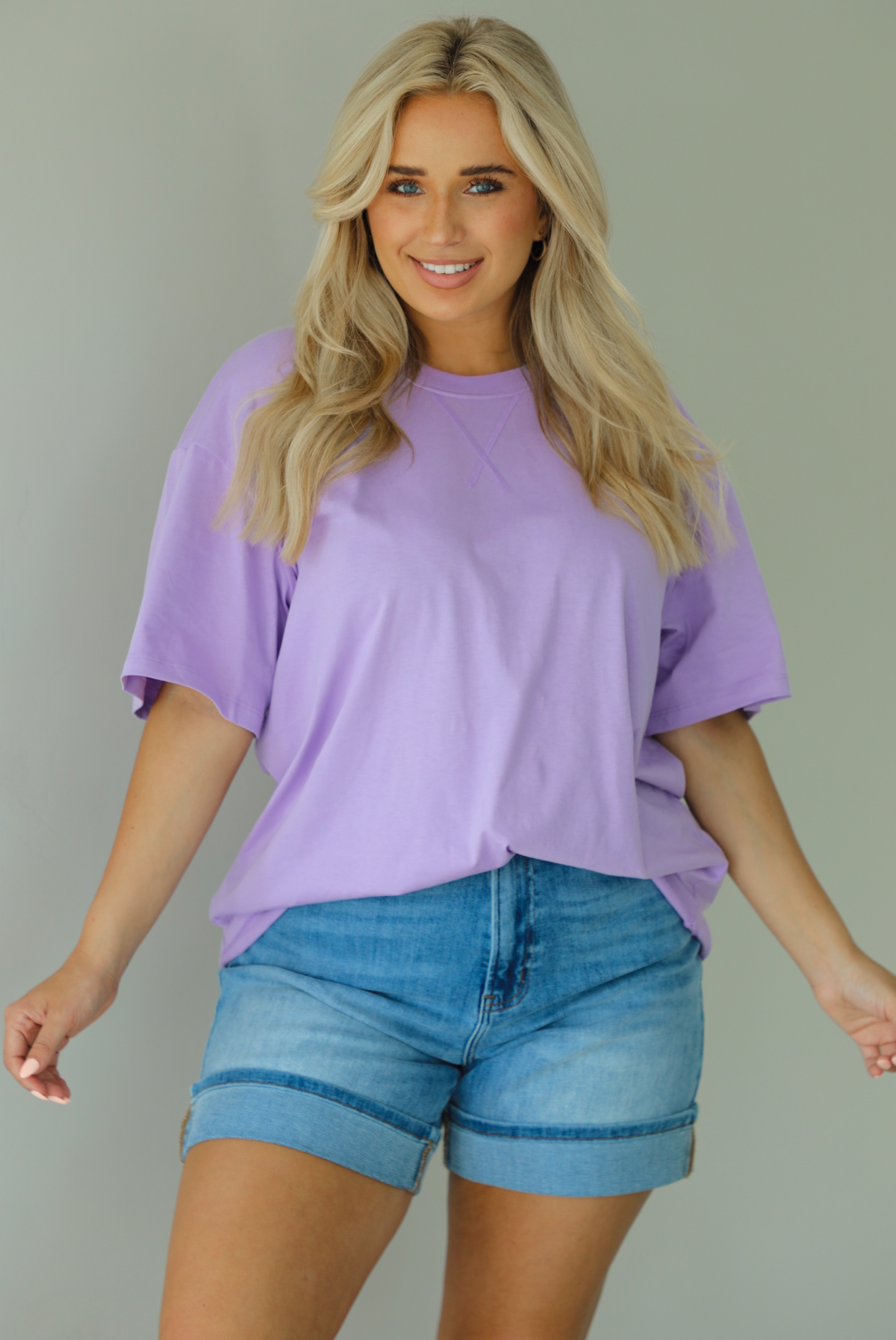 Woman wearing a lavender oversized tee tucked into medium wash denim shorts indoors against a white wall.