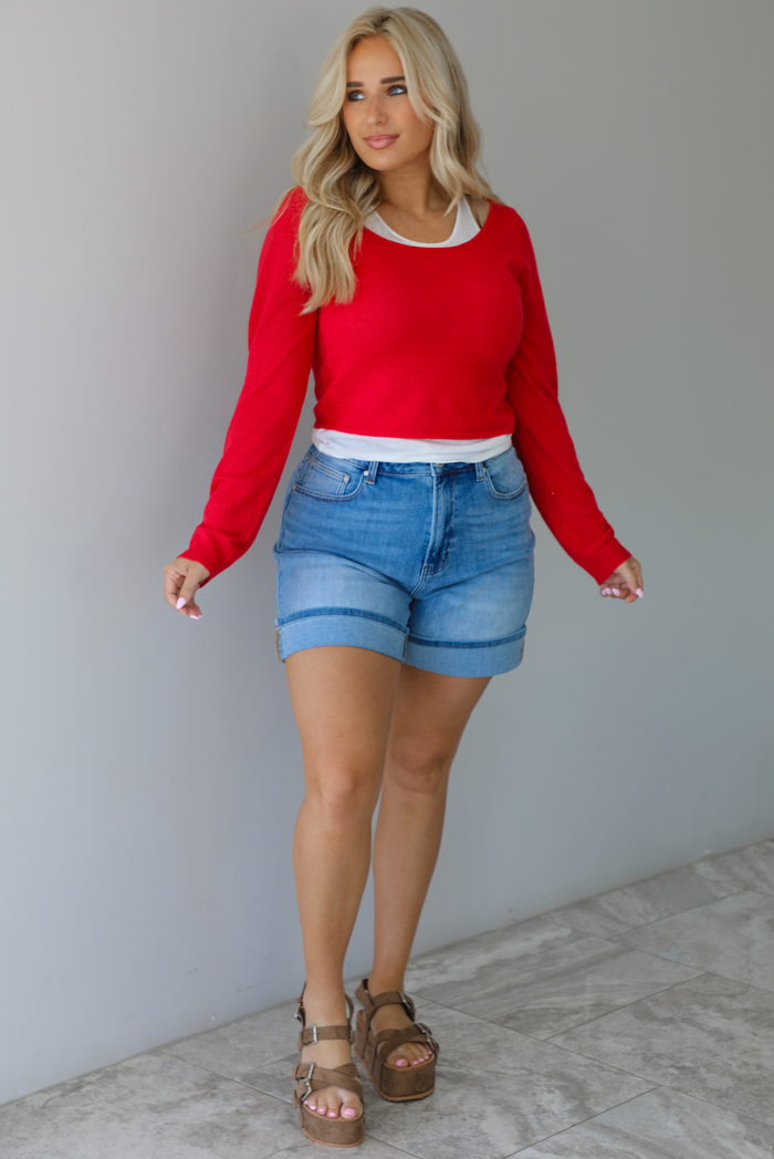 Woman wearing a red, long sleeved knit top with a white tank underneath with medium wash denim shorts indoors against a white wall with white marble flooring. 