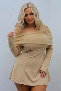 Woman wearing a tan mini dress that has a folded over detail at the shoulders that is a ribbed textured knit material, with a soft, stretchy remainder of the dress with long sleeved, and faux-button details on the front in the shape of a square. The photo is taken outdoors against a white wall. 