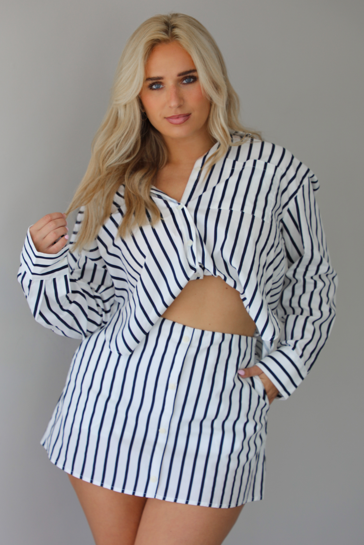 Woman wearing a white and navy striped set with a collared button-down top and mini skirt indoors against a white wall.