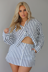 Woman wearing a white and navy striped set with a collared button-down top and mini skirt indoors against a white wall.