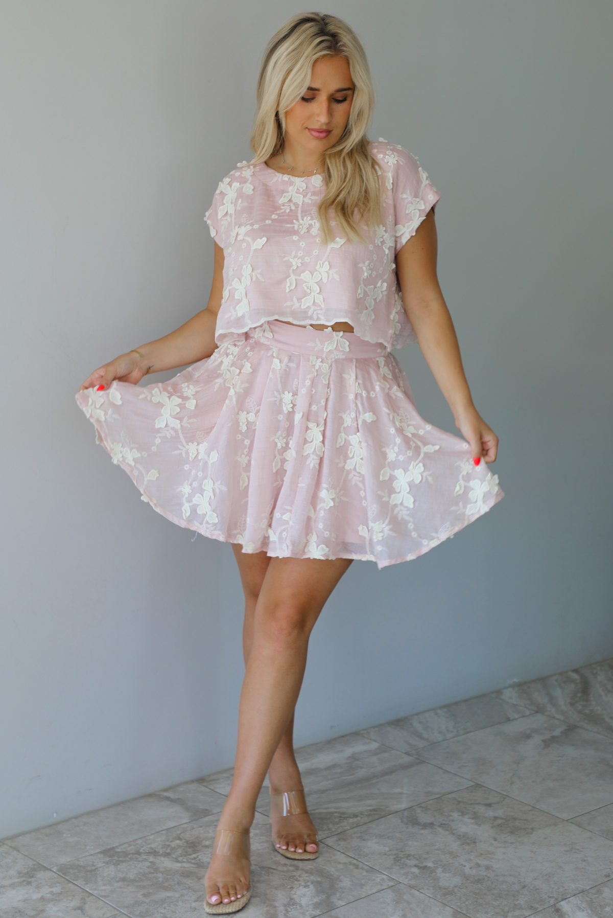 Woman wearing a pink and white floral embroidered two piece set featuring a cropped loose fit top and high waisted skirt indoors against a white wall with white marble flooring.