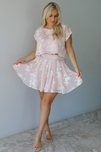 Woman wearing a pink and white floral embroidered two piece set featuring a cropped loose fit top and high waisted skirt indoors against a white wall with white marble flooring.