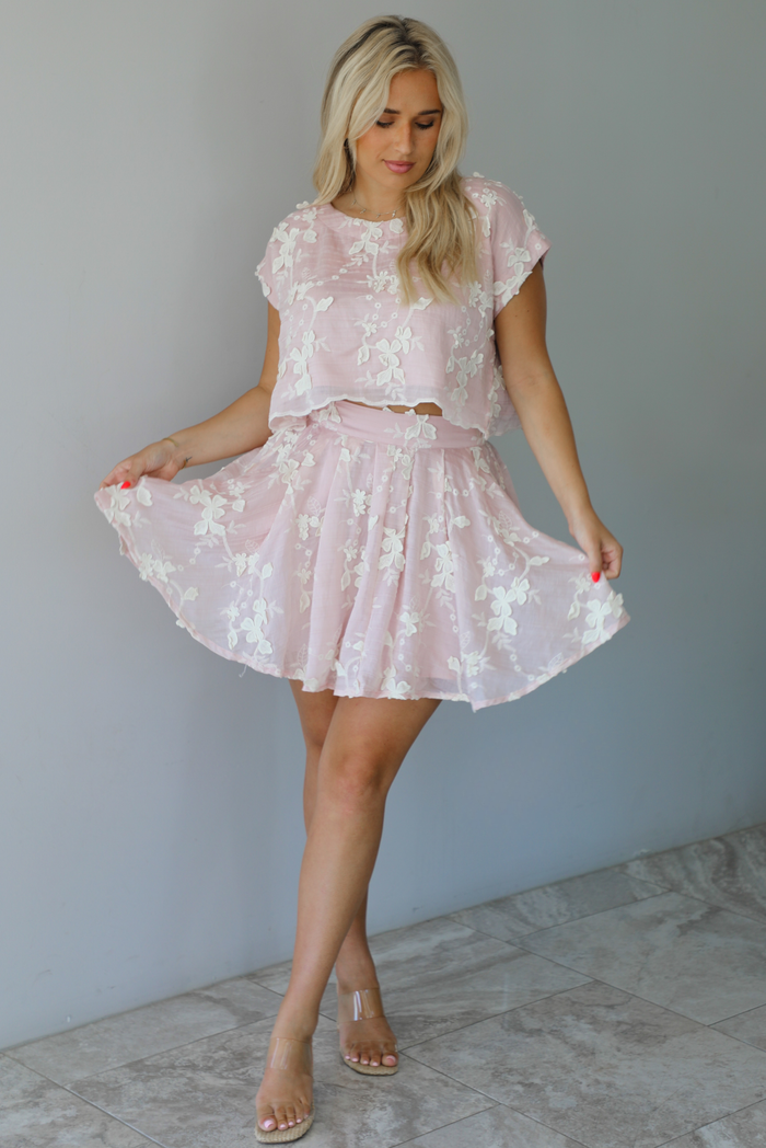 Woman wearing a pink and white floral embroidered two piece set featuring a cropped loose fit top and high waisted skirt indoors against a white wall with white marble flooring.