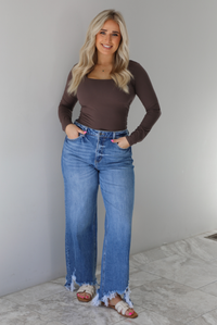 Woman wearing a brown long sleeved fitted top tucked into high waisted distressed denim. The photo is taken indoors against a white wall with white marble flooring. 