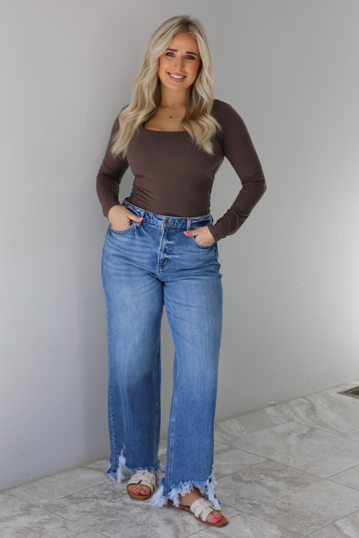 Woman wearing a brown long sleeved fitted top tucked into high waisted distressed denim. The photo is taken indoors against a white wall with white marble flooring. 