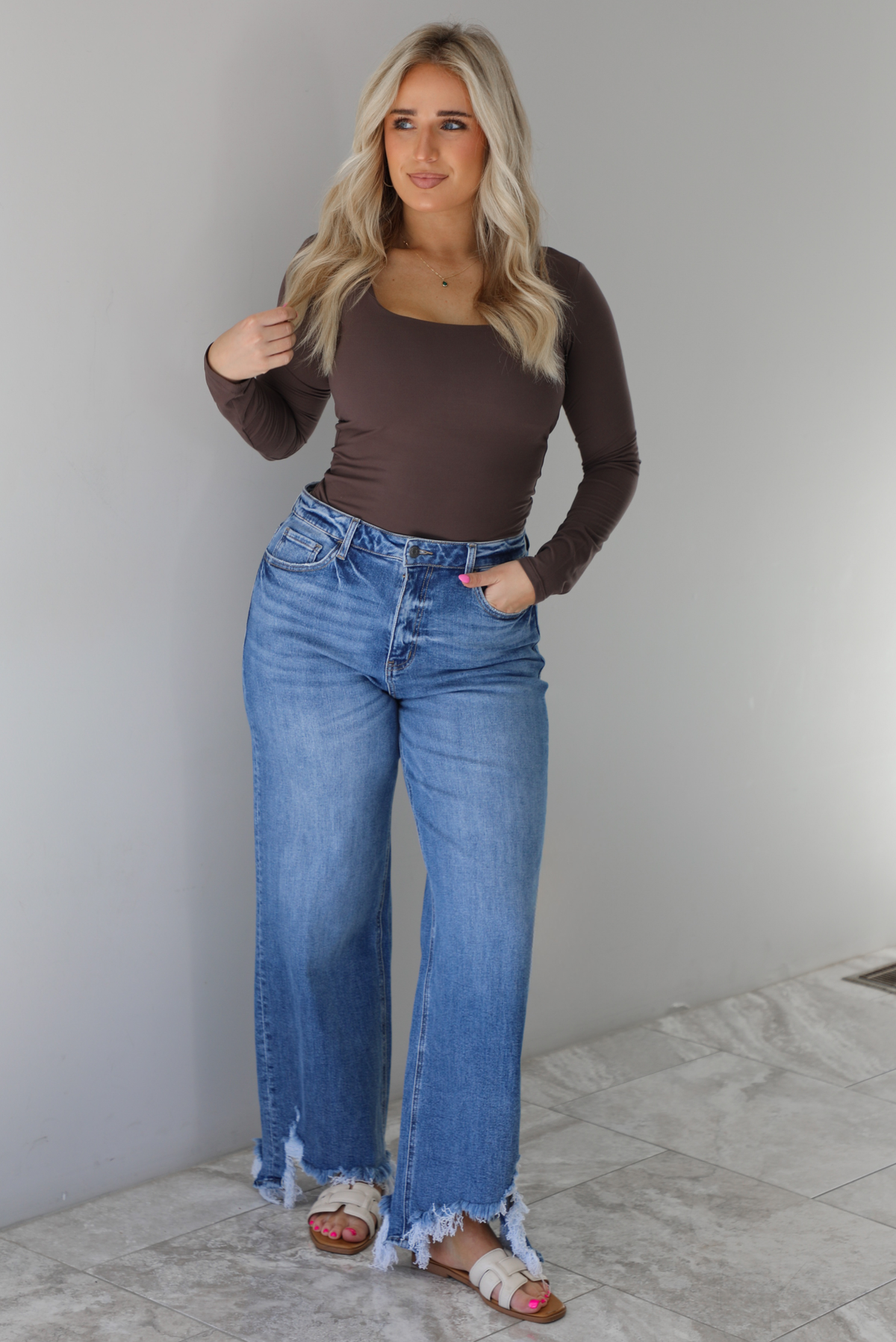 Woman wearing a brown long sleeved fitted top tucked into high waisted distressed denim. The photo is taken indoors against a white wall with white marble flooring. 