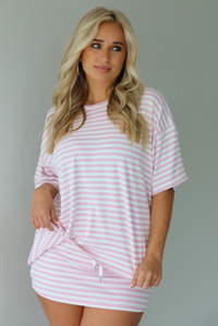 Woman wearing a pink and white striped two piece set featuring an oversized two and fitted mini skirt indoors against a white wall. 