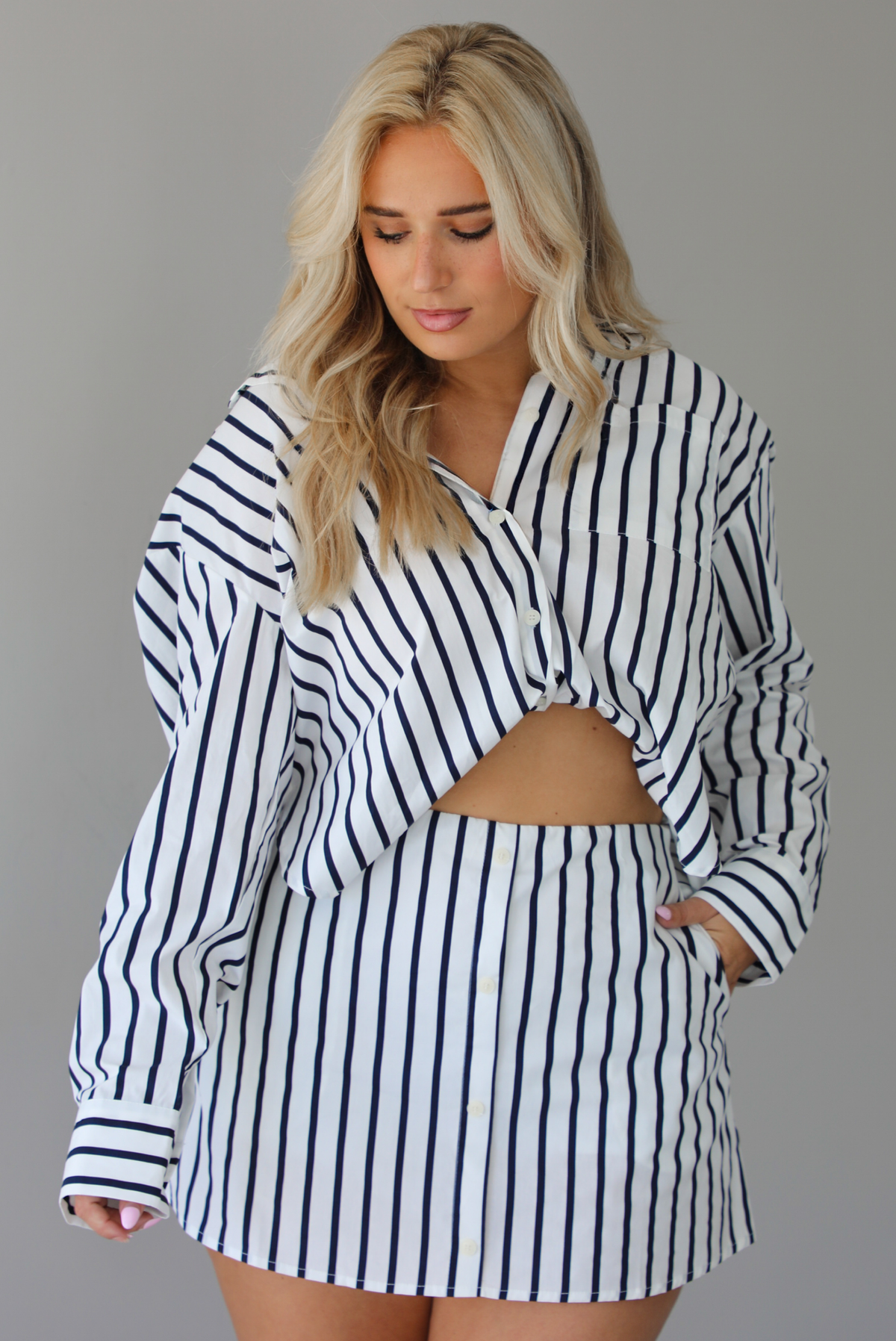 Woman wearing a white and navy striped set with a collared button-down top and mini skirt indoors against a white wall.
