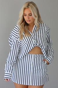 Woman wearing a white and navy striped set with a collared button-down top and mini skirt indoors against a white wall.