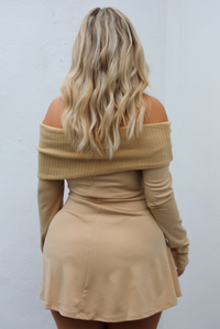 Woman wearing a tan mini dress that has a folded over detail at the shoulders that is a ribbed textured knit material, with a soft, stretchy remainder of the dress with long sleeved, and faux-button details on the front in the shape of a square. The photo is taken outdoors against a white wall.  The photo shows the back of the dress, which looks like the front, but doesn't have the button details. 