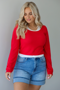Woman wearing a red, long sleeved knit top with a white tank underneath with medium wash denim shorts indoors against a white wall.