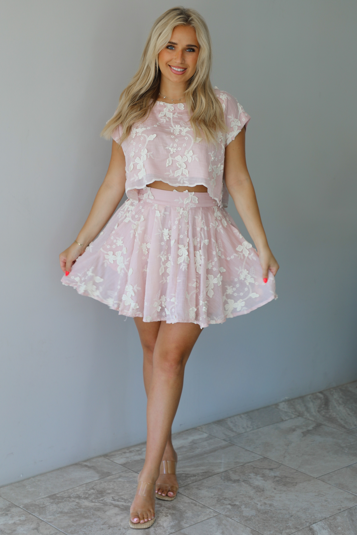 Woman wearing a pink and white floral embroidered two piece set featuring a cropped loose fit top and high waisted skirt indoors against a white wall with white marble flooring.