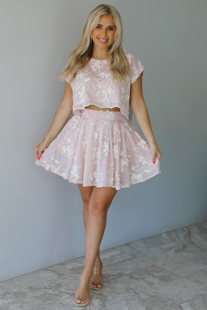 Woman wearing a pink and white floral embroidered two piece set featuring a cropped loose fit top and high waisted skirt indoors against a white wall with white marble flooring.