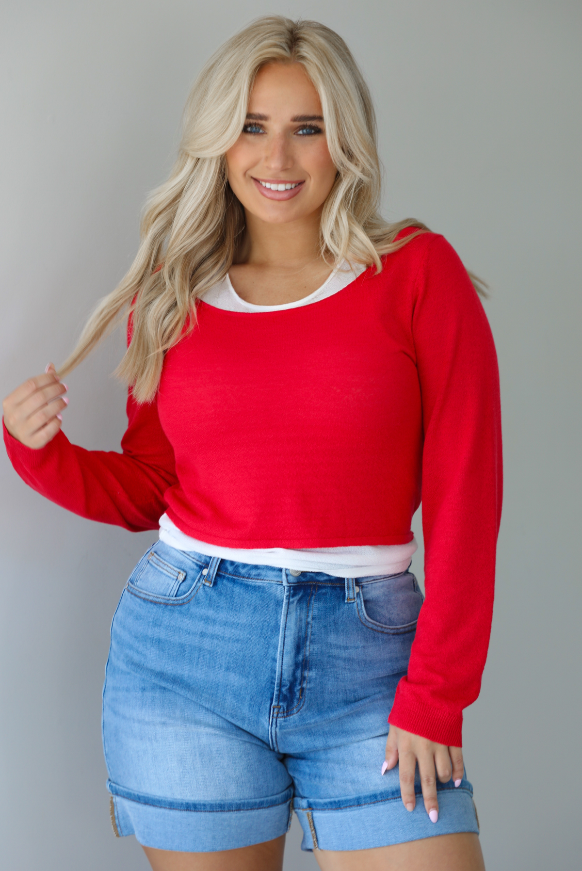 Woman wearing a red, long sleeved knit top with a white tank underneath with medium wash denim shorts indoors against a white wall.