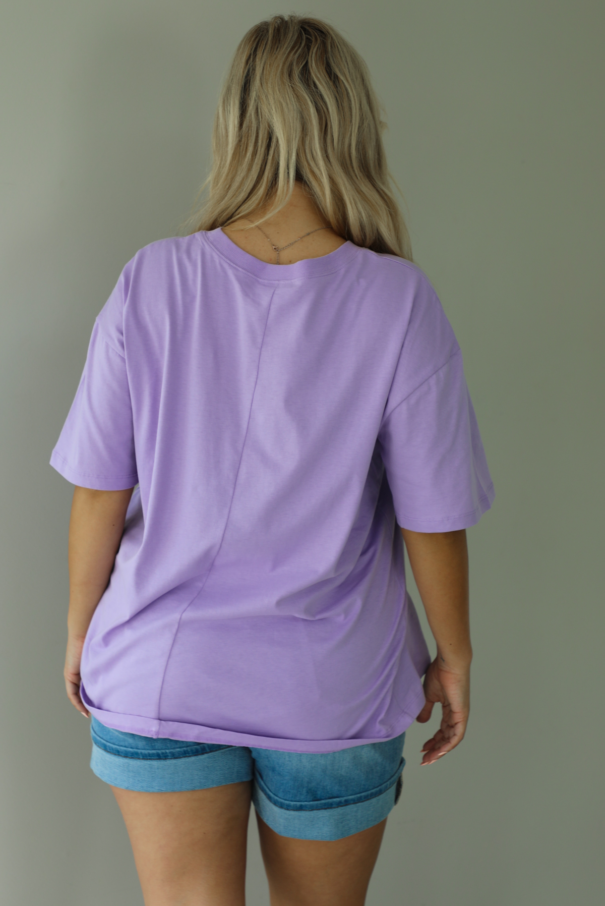 Woman wearing a lavender oversized tee untucked with medium wash denim shorts indoors against a white wall. The photo shows the back of the tee. 