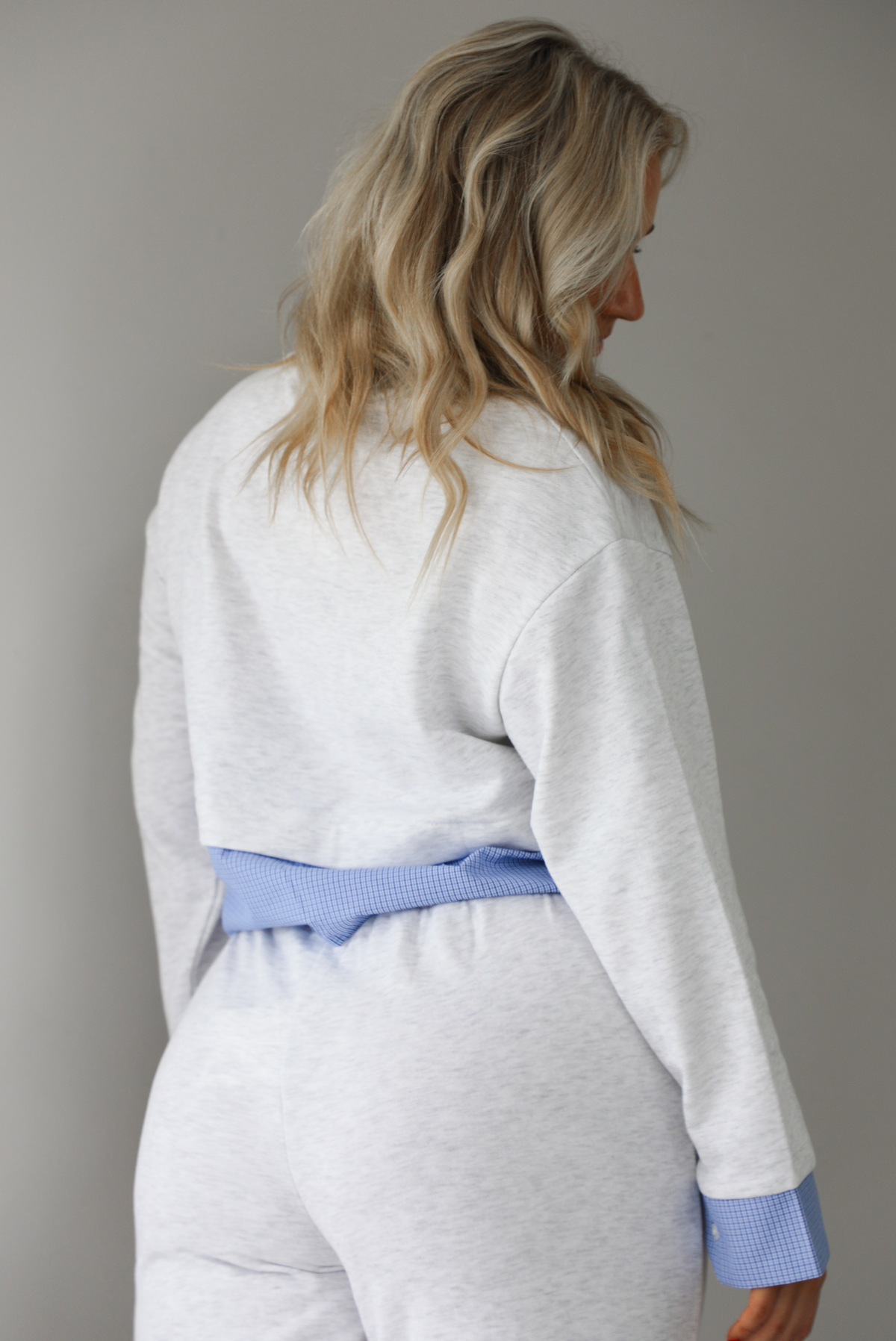 Woman wearing a heather grey two piece set with blue and white pin striped detailing at the hem of the top, at the wrists on the top, and at the waistline on the pants. The photo shows a closeup of the back of the top and upper portion of the pants. The photo is taken indoors against a white wall with white marble flooring. 