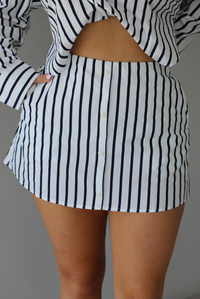 Woman wearing a white and navy striped set with a collared button-down top and mini skirt indoors against a white wall and shows a closeup of the skirt, which has functional pockets on either side and non-functional button closures in the front. 