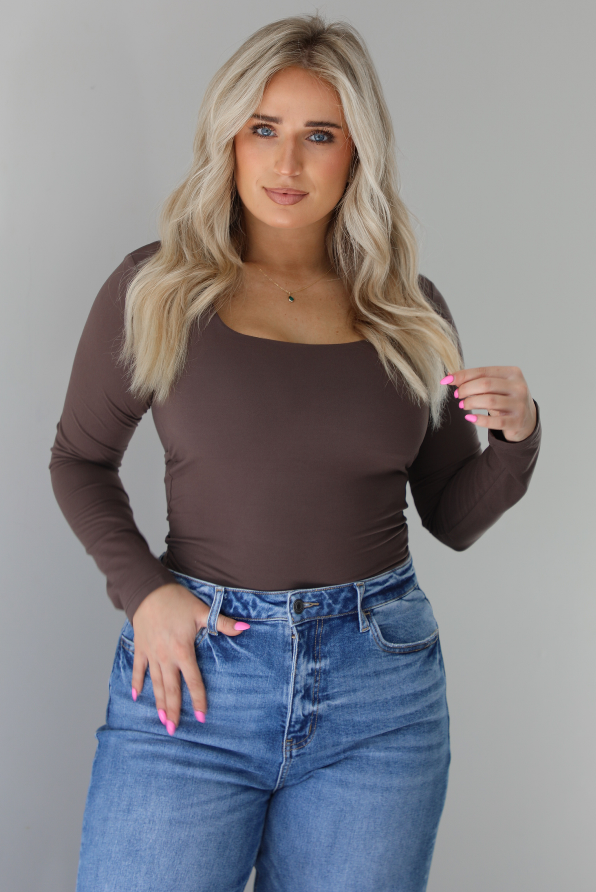 Woman wearing a brown long sleeved fitted top tucked into high waisted distressed denim. The photo is taken indoors against a white wall.