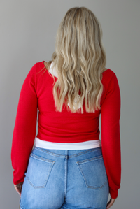 Woman wearing a red, long sleeved knit top with a white tank underneath with medium wash denim shorts indoors against a white wall and shows the back of the top. 