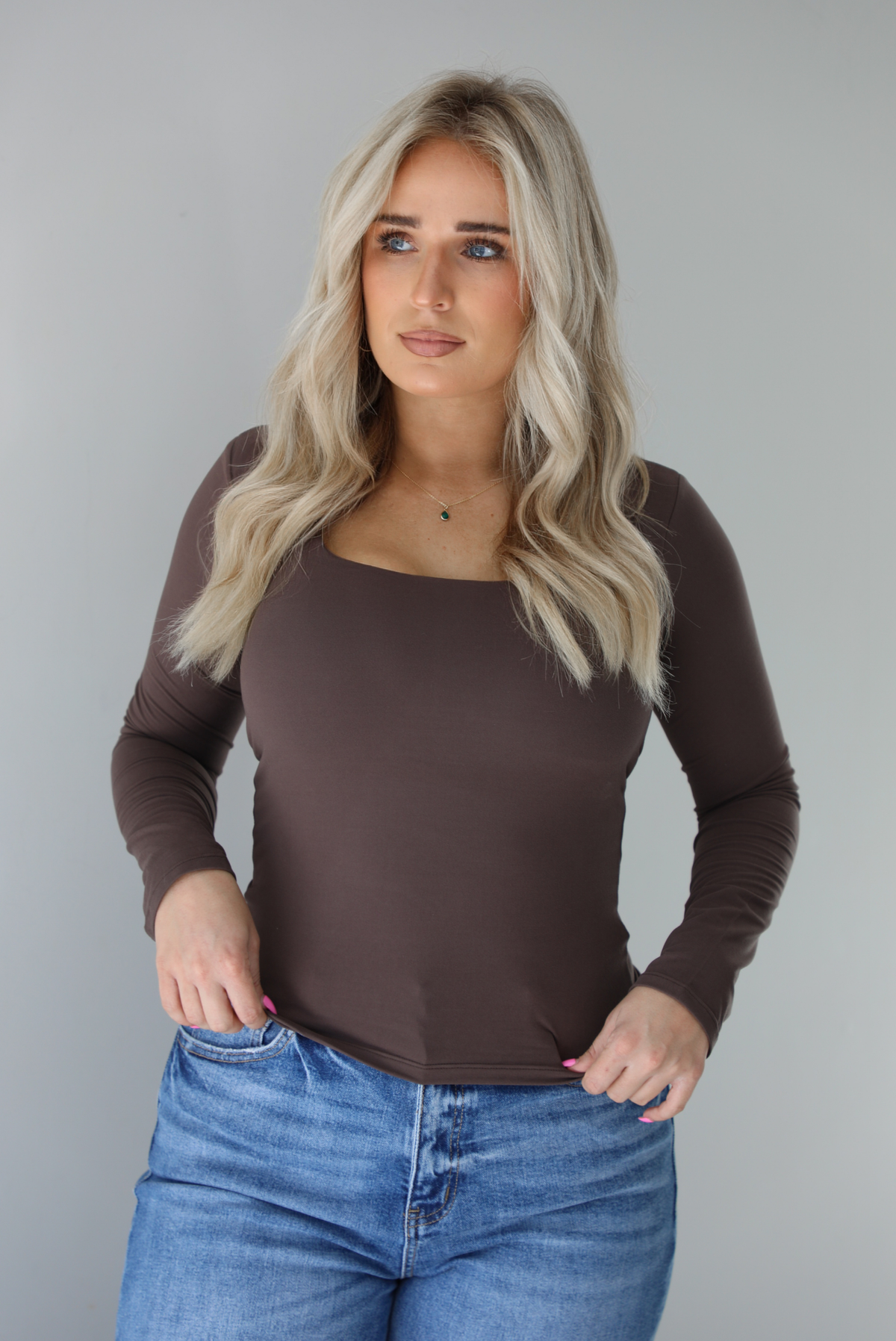 Woman wearing a brown long sleeved fitted top untucked from high waisted distressed denim. The photo is taken indoors against a white wall.