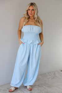 Woman wearing a blue two piece set featuring a strapless smocked top with a ruffled hem and high waisted flared pants with functional pockets on either side. The photo is taken indoors against a white wall with white marble flooring. 