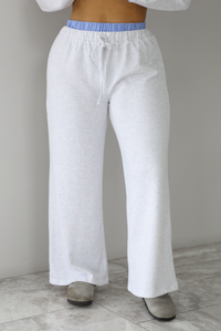 Woman wearing a heather grey two piece set with blue and white pin striped detailing at the hem of the top, at the wrists on the top, and at the waistline on the pants. The photo shows a closeup of the full length of the front of the pants. The photo is taken indoors against a white wall with white marble flooring. 