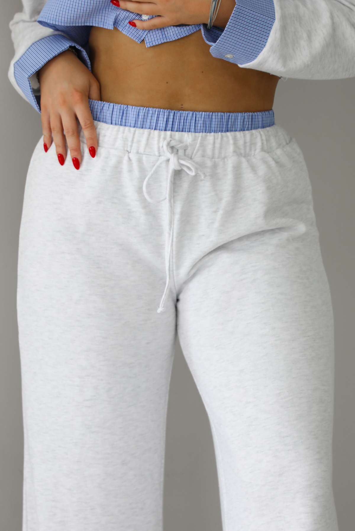 Woman wearing a heather grey two piece set with blue and white pin striped detailing at the hem of the top, at the wrists on the top, and at the waistline on the pants. The photo shows a closeup of the waistline of the pants..The photo is taken indoors against a white wall with white marble flooring. 