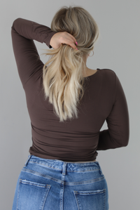 Woman wearing a brown long sleeved fitted top untucked from high waisted distressed denim. The photo shows the back of the top. The photo is taken indoors against a white wall.