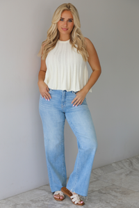 Woman wearing a high neck cream flowy knit top with light-wash denim jeans in front of a white wall with white marble flooring. 
